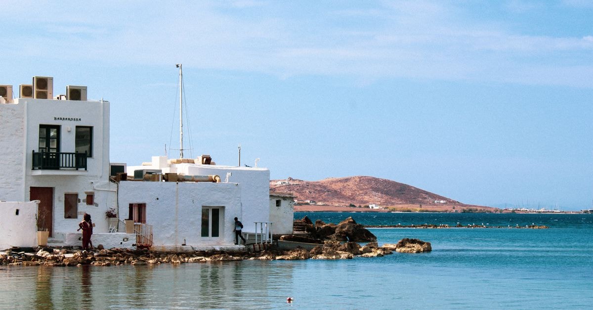 Ermoupoli Travel Guide: Cycladic Charm in Syros's Harbor Capital Charming white buildings by the coast in Naoussa, Paros Island with serene blue waters and hills in the background.