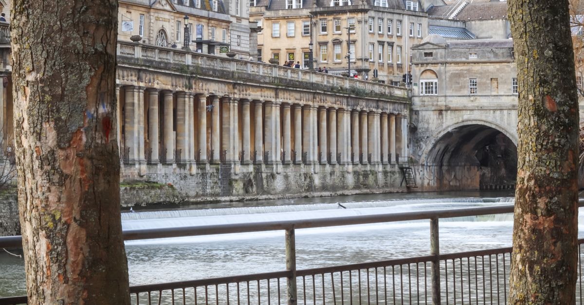Bath Travel Guide: Planning Tips, Costs and Things to Do Elegant architectural buildings along a river in a historic European city, framed by trees.