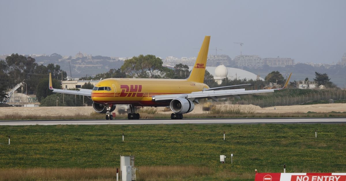 晴朗的日子裡，一架 DHL 貨機在機場跑道上滑行，背景為城市天際線。