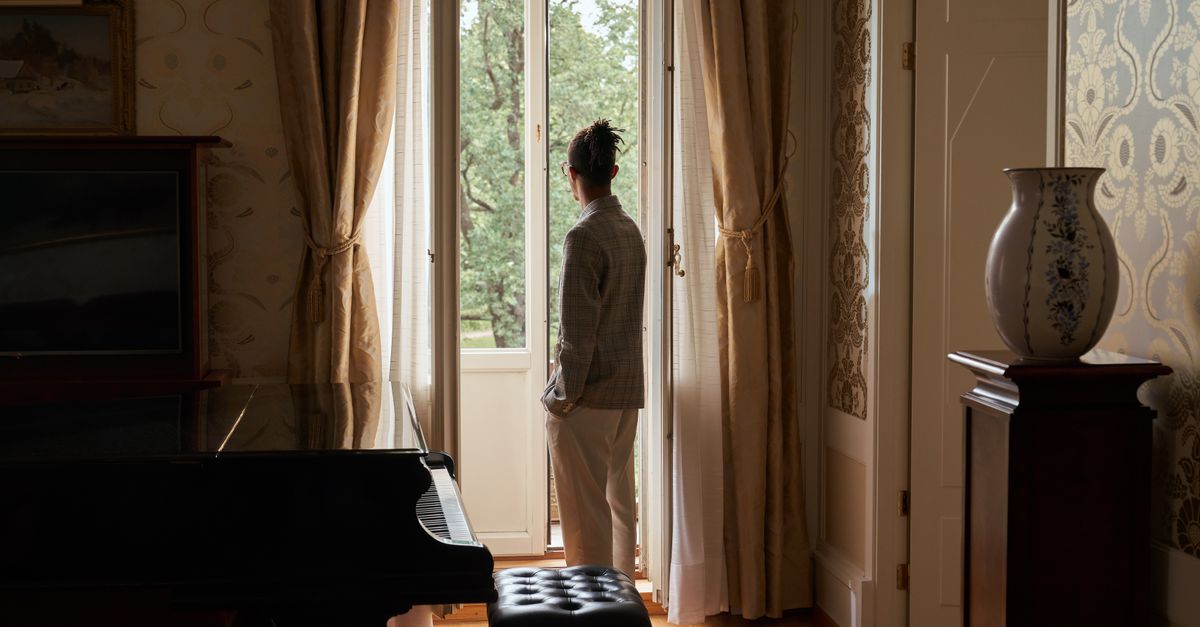 A man stands in an elegant room, looking out the window with a grand piano nearby.