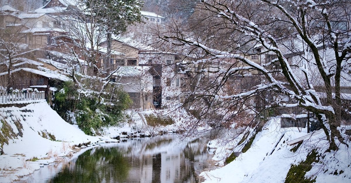 A picturesque winter view of snow-covered trees and traditional houses by a river in Takayama, Japan.