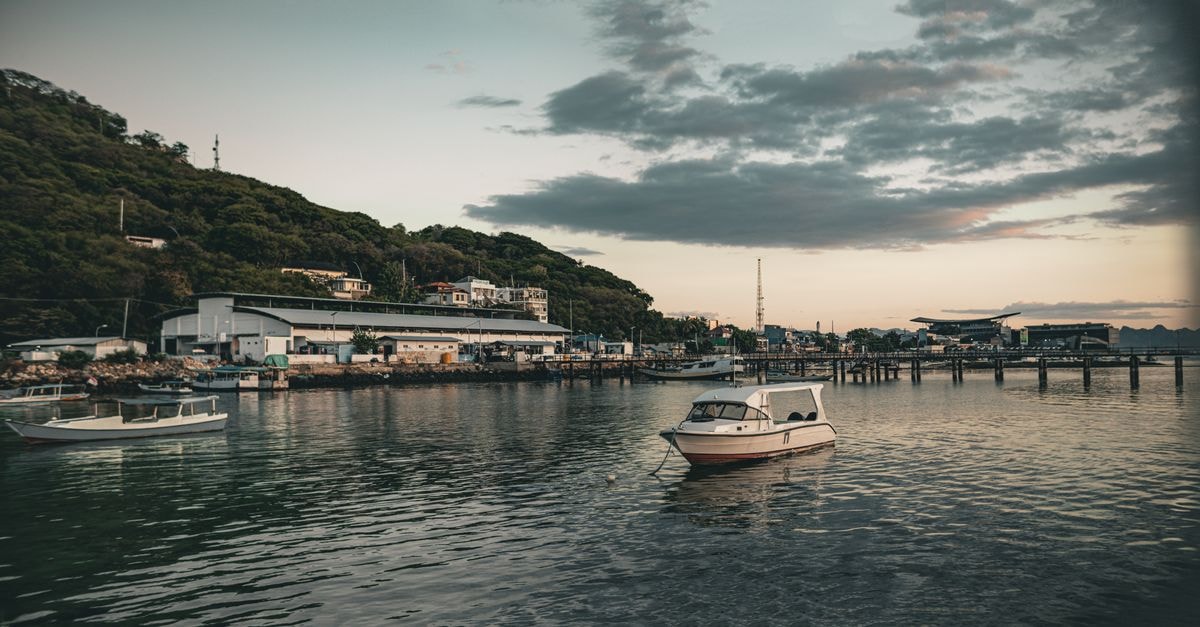 Serene coastal landscape with boats anchored in a harbor during sunset, calm waters and hills.