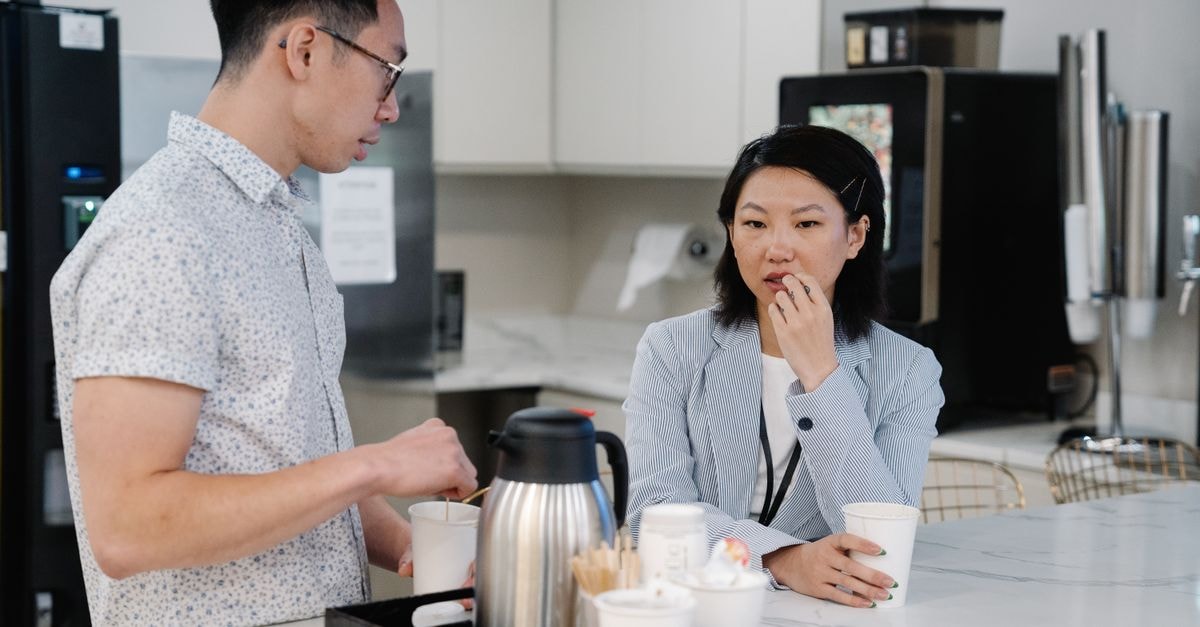 Two colleagues discussing over coffee in a modern office break room setting.