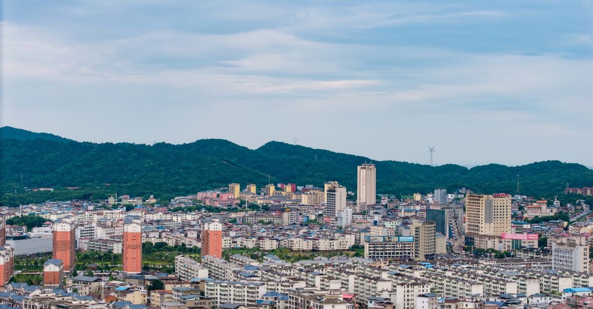 Aerial view of a cityscape with tall buildings surrounded by lush green hills under a cloudy sky.