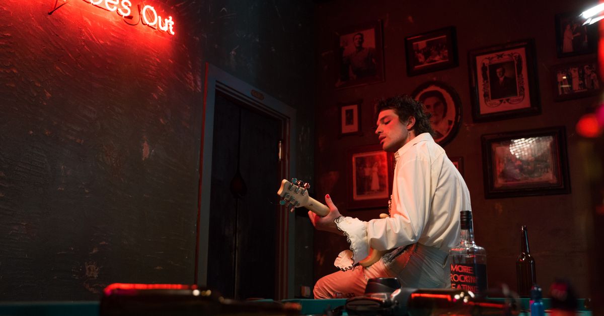 Green Day Unveils 'I'm Never Gonna R.I.P.' from NIMRODS Movie Soundtrack A musician plays electric guitar in a bar under neon lights, creating a punk atmosphere.