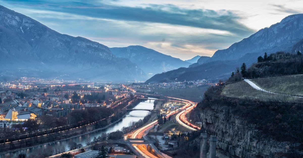 A Slow Travel Guide to Trento: Getting Around and Eating Well A stunning aerial view of Trento, Italy at twilight with mountains, highways, and a river.