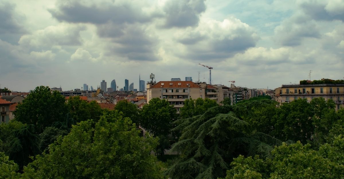 Room Mate Giulia Milan: Patricia Urquiola–Designed Boutique Hotel Near the Duomo Skyline of Milan with clear view of urban buildings amidst lush green trees under a cloudy sky.