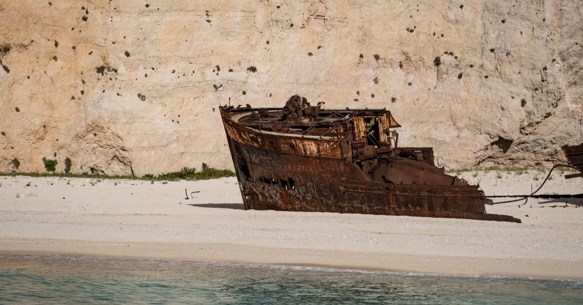 A rusty shipwreck rests on a serene sandy beach against a rocky cliff, evoking adventure and travel.