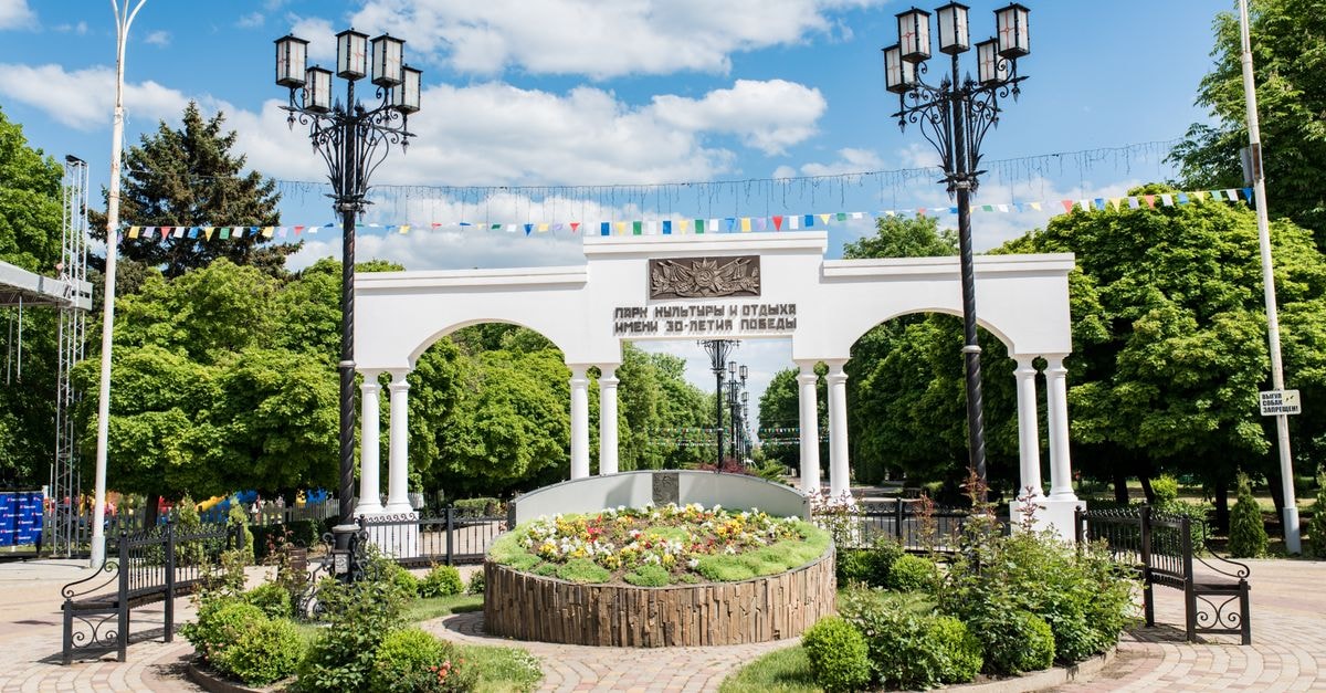 Krasnodar Travel Guide: Getting Around and Eating Well Elegant white archway and vibrant garden at a city park entrance on a sunny day.