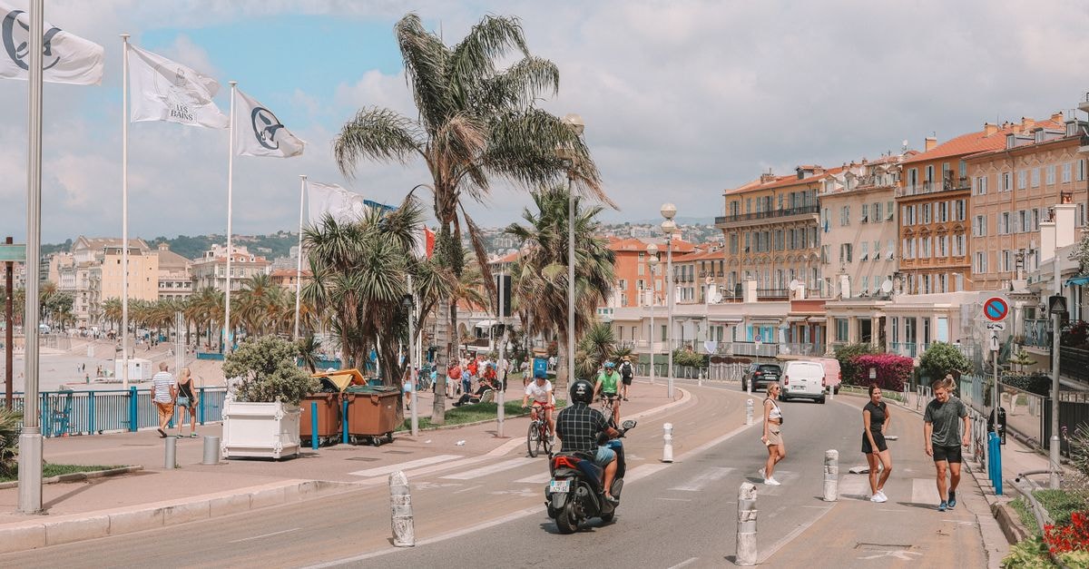 Pedestrians and scooters on bustling Promenade des Anglais in Nice, France, with palm trees and coastal buildings.