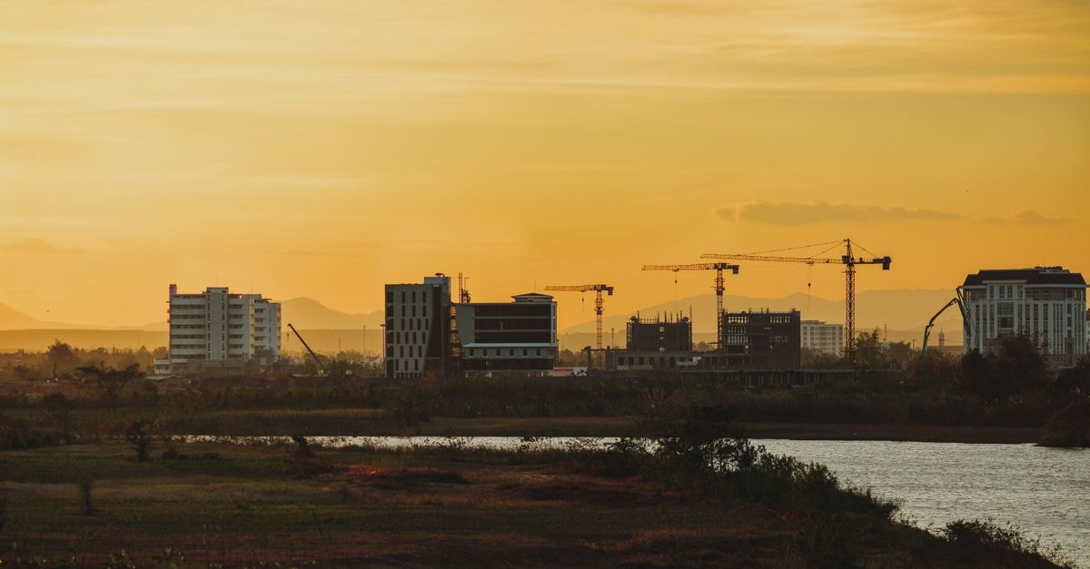 A vibrant sunset view of the Kon Tum city skyline with construction cranes and buildings in silhouette.