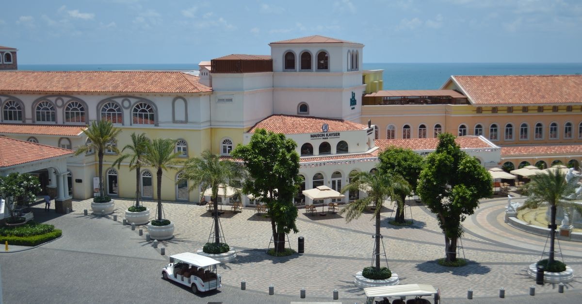 Haikou Travel Guide: Getting Around and Eating Well Aerial view of a Mediterranean-style plaza with buildings and palm trees by the ocean, featuring clear skies.