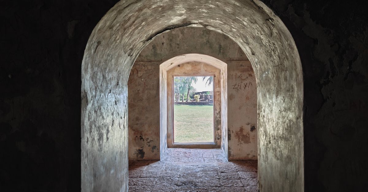 Stone archway with sunlight highlighting rough textures, leading to a green garden. Captured in Kannur, India.