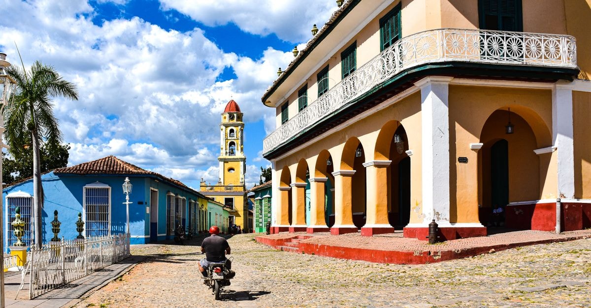 A motorcyclist rides down sunlit cobblestone streets in a colorful Cuban town, showcasing colonial architecture.