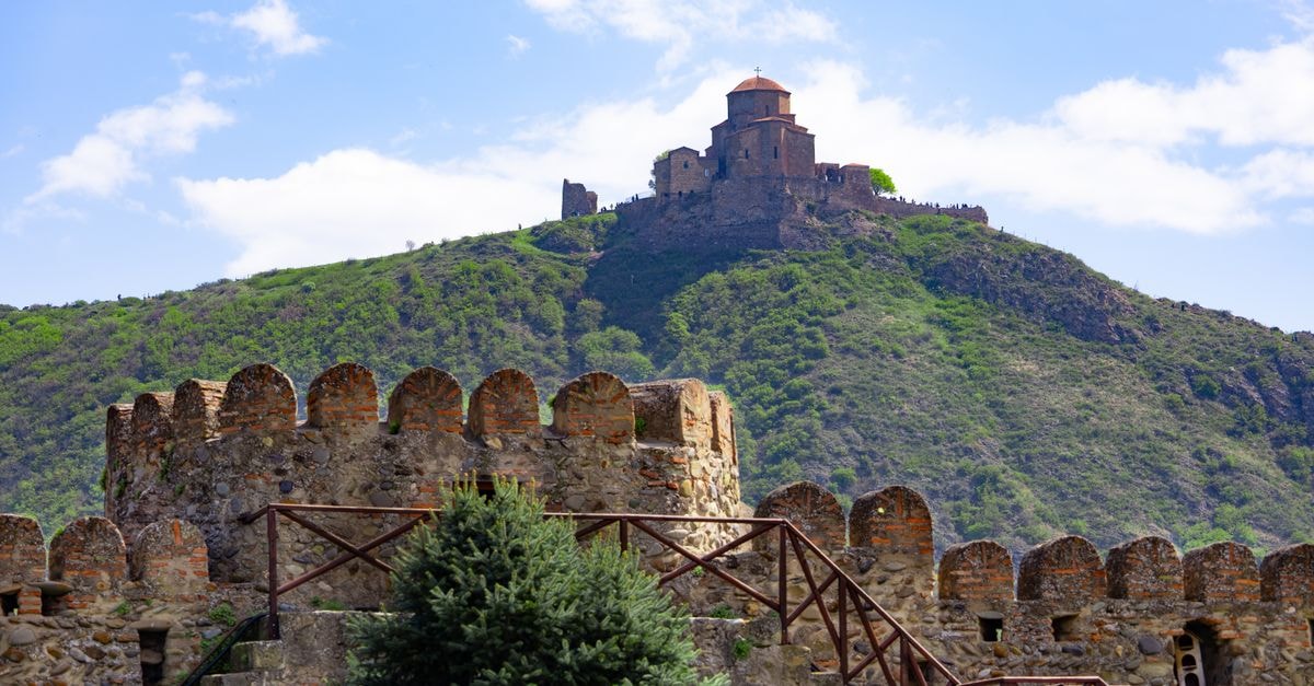 Jvari Monastery atop a green hill under blue sky in Mtskheta, Georgia, with foreground fortress.