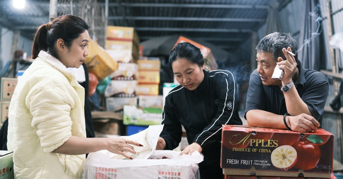Three adults working together in a Vietnamese warehouse, engaged in discussion amid apple boxes.