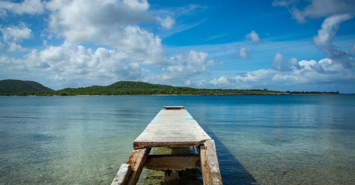 Serene ocean view with a rustic pier under a vibrant blue sky and lush island in the background.
