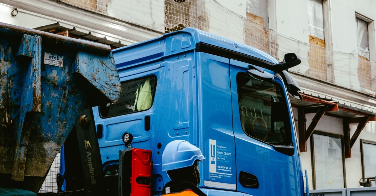 Caterpillar at a Cyclically Stretched Multiple Construction worker standing by a blue truck at an urban building site, wearing a safety vest and hard hat.