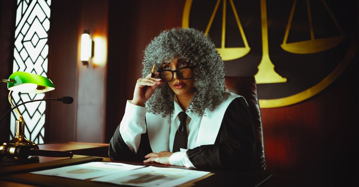 Judge in traditional attire sits at a courtroom desk with a serious expression.