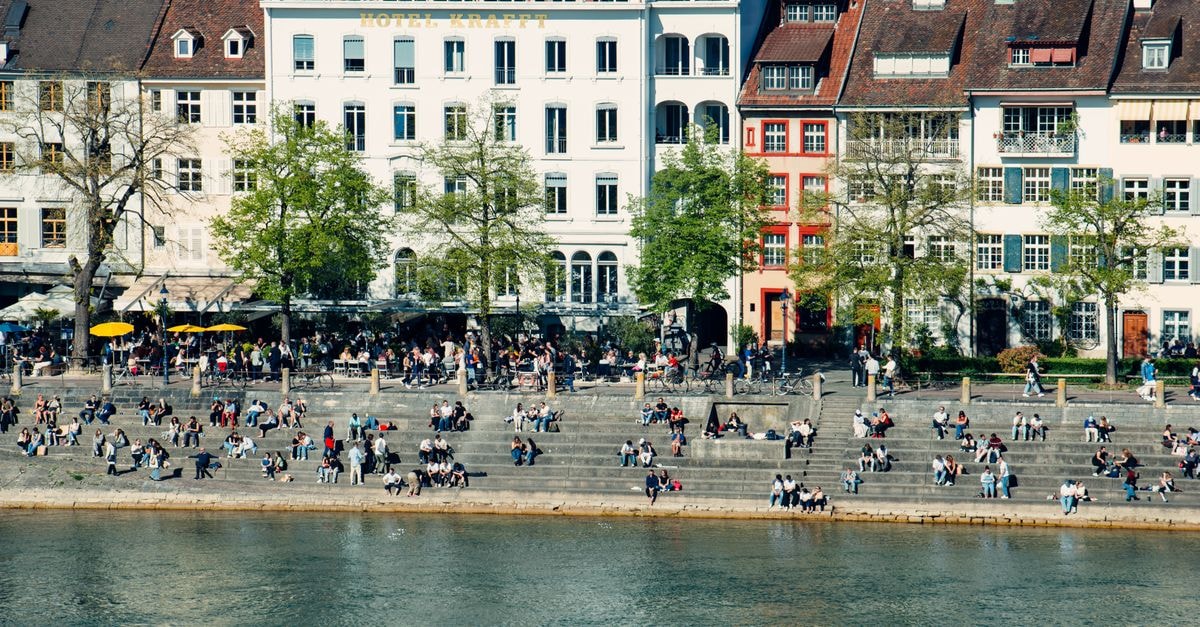 A Quietly Magnetic Basel Travel Guide People relax and socialize by the Rhine River in Basel, showcasing vibrant city life.
