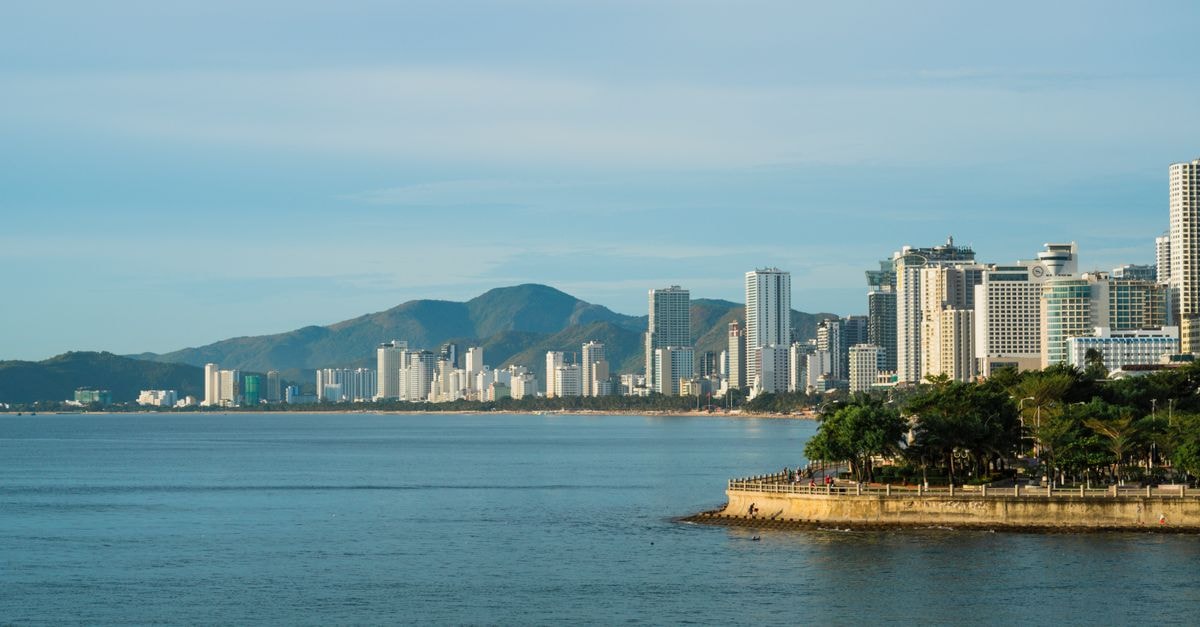 A serene view of a modern coastal city skyline against majestic mountains under a clear blue sky.