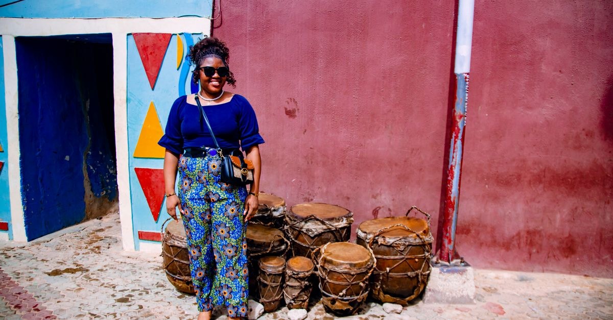 Smiling woman in sunglasses beside traditional drums and vibrant murals. Urban setting.