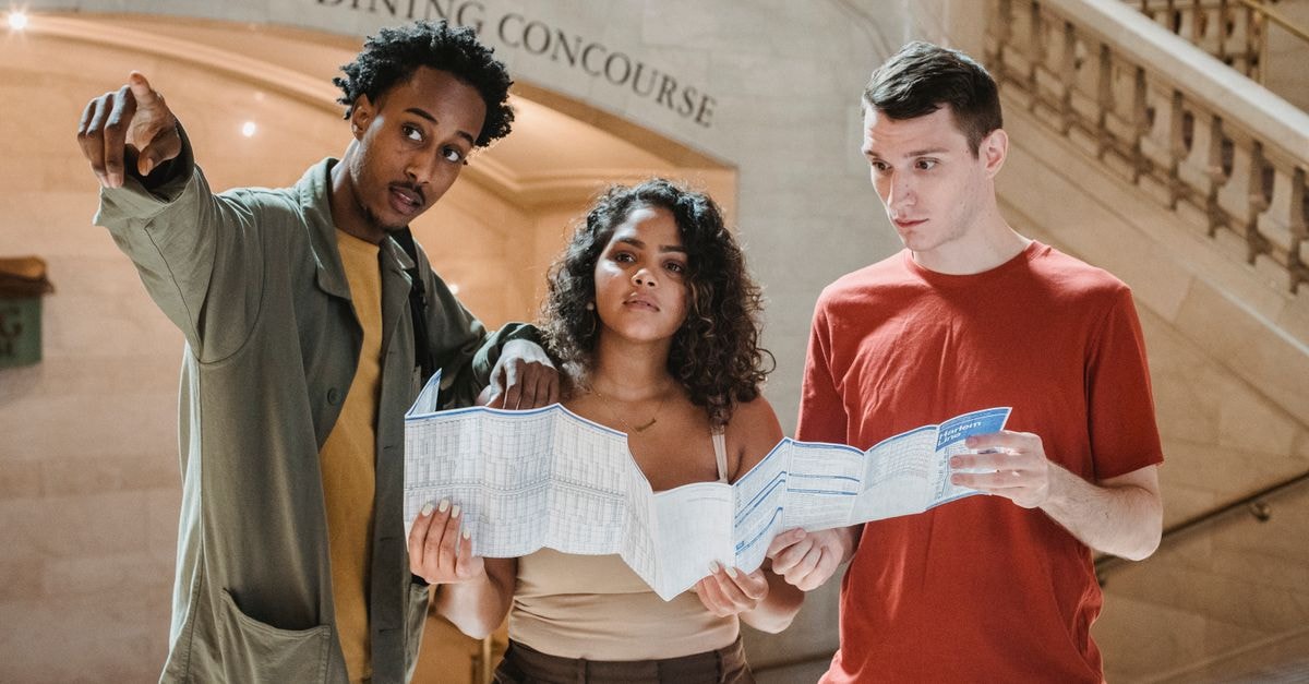 Kolwezi Travel Guide: How to Plan a Practical Visit Young African American male tourist pointing away while searching for direction with diverse fiends standing in railway station terminal with paper map in hands