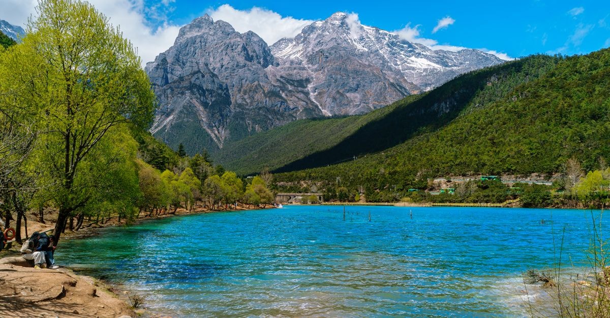 Liupanshui Travel Guide: China's Cool City for Forest Air and Easy Days Scenic view of a snowy mountain by a tranquil blue lake, lush trees around.