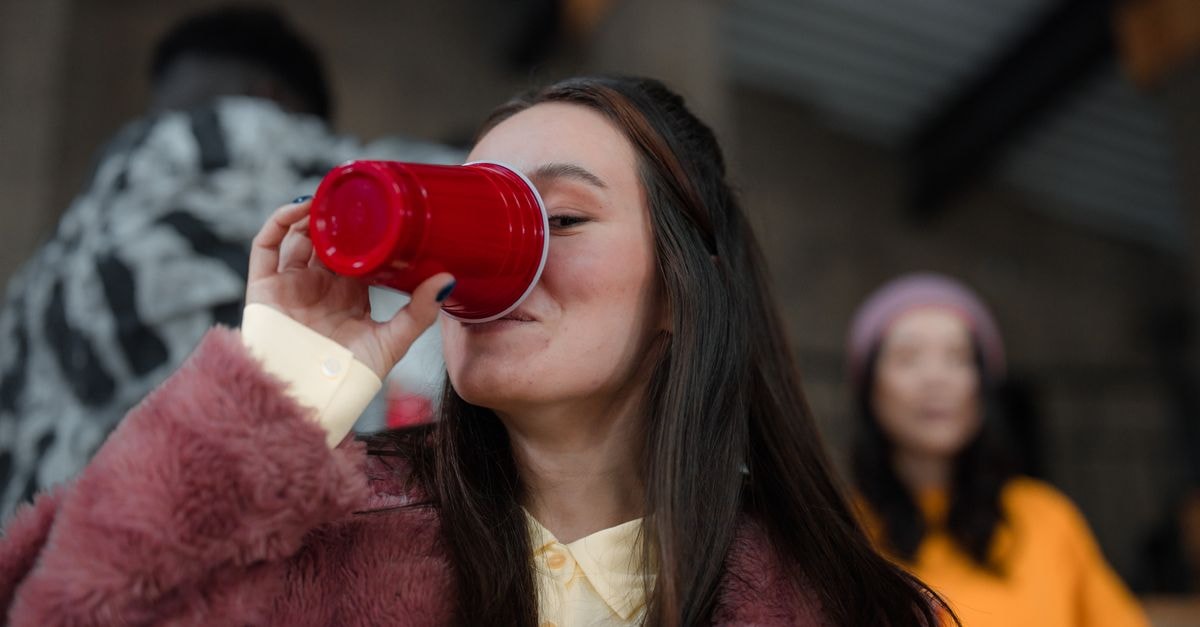 A stylish young woman drinks from a red cup during a lively indoor gathering.