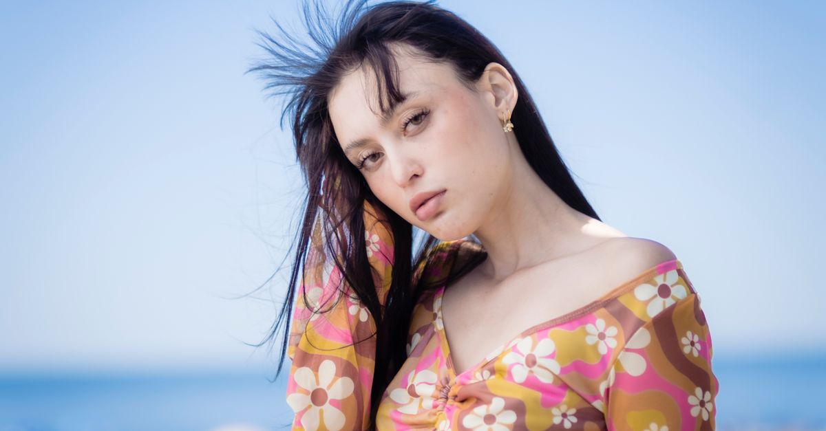 Gracie Abrams Tops Billboard 200 for the First Time With 'Daughter From Hell' A fashionable woman wearing a floral top poses on a sunny beach with the ocean in the background.