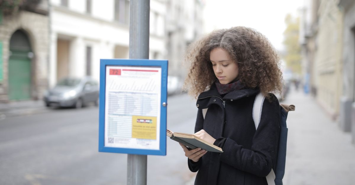 Awka Travel Guide: Planning a Considered City Stop A young woman with curly hair reads a book while standing at a bus stop on a city street.