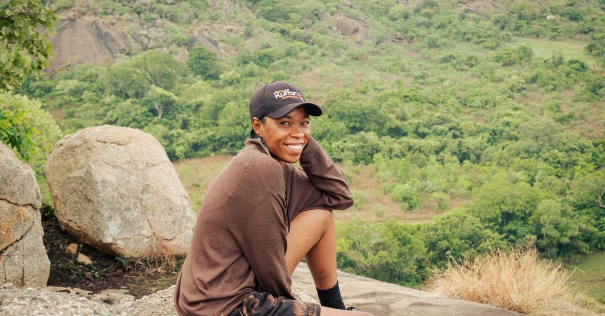 A cheerful woman enjoys a peaceful moment sitting on a rocky mountain with a lush green view.