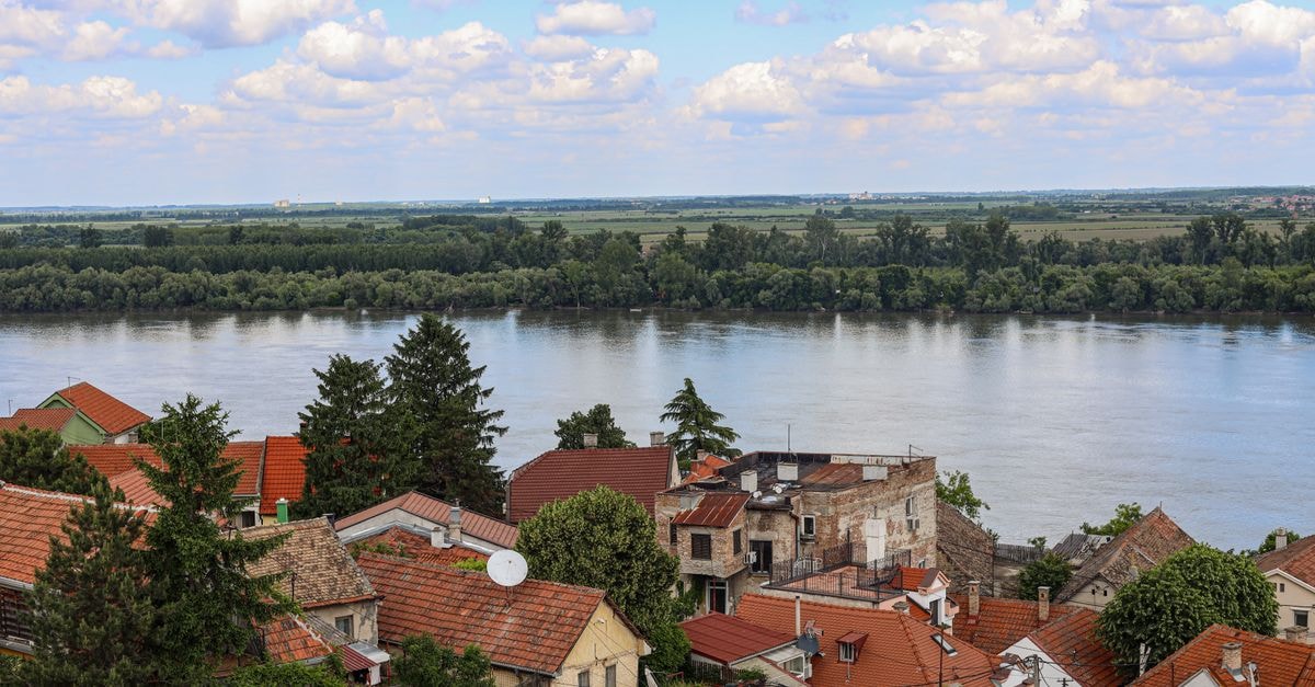 Charming riverside town with red-roofed houses and lush greenery under a blue sky.