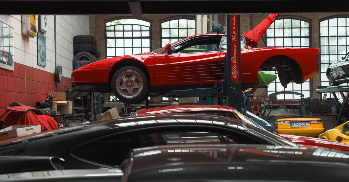 A vibrant red sports car is elevated on a lift inside an auto repair garage, surrounded by various vehicles and equipment.