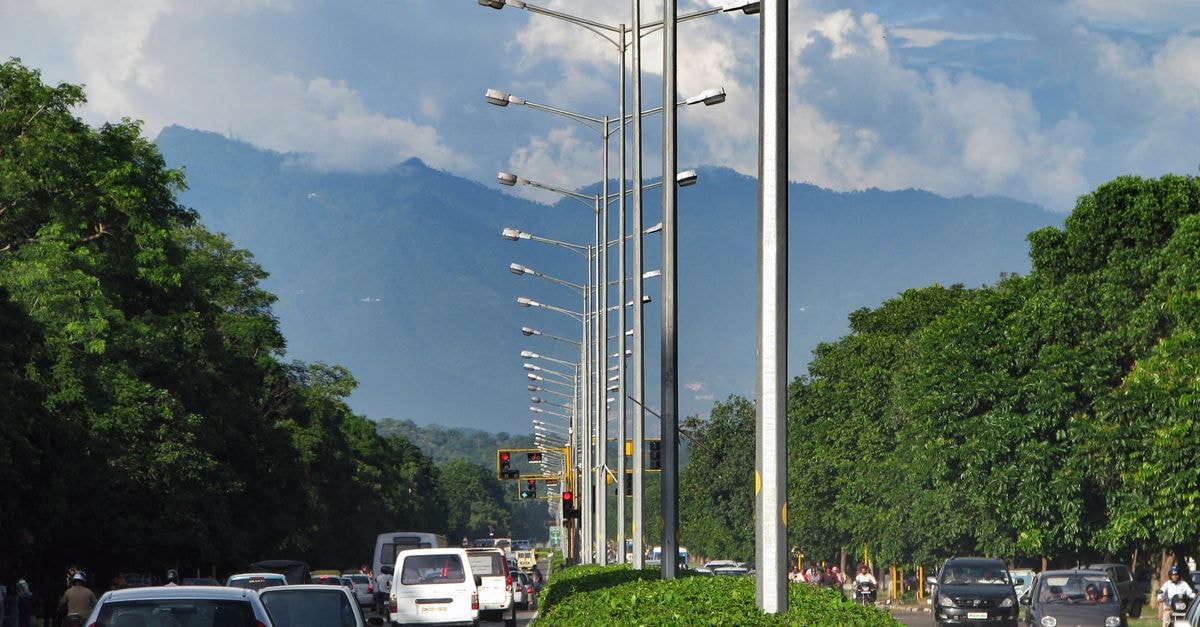 SWAGATAM INN, Guwahati — A 5-Minute Walk From The Railway Station Traffic flows on a tree-lined road with stunning mountain backdrop.