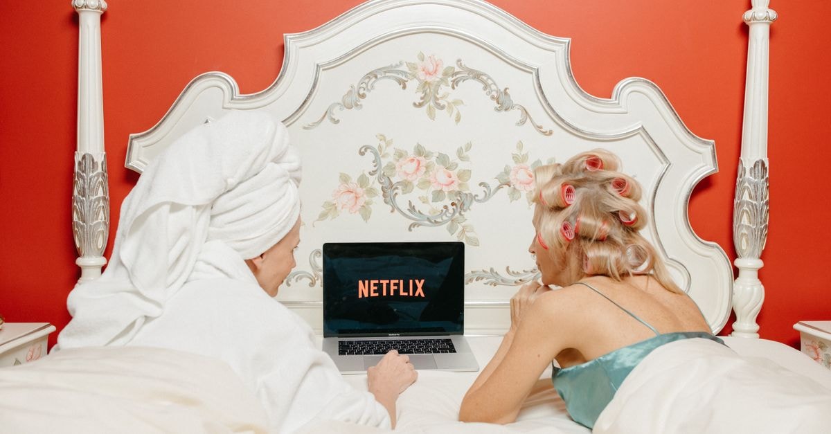 HBO Max's 'Lanterns' Premieres Sunday With Simultaneous HBO Linear Drop Two women enjoying Netflix in a cozy, vintage-style bedroom with red walls.