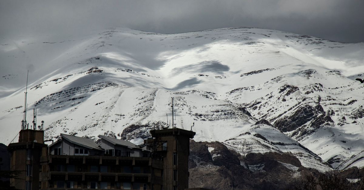 Rooberoo Hostel Tehran: A Cultural Hub Stay in the Heart of Tehran Snow-covered mountains loom over urban buildings under a moody sky.