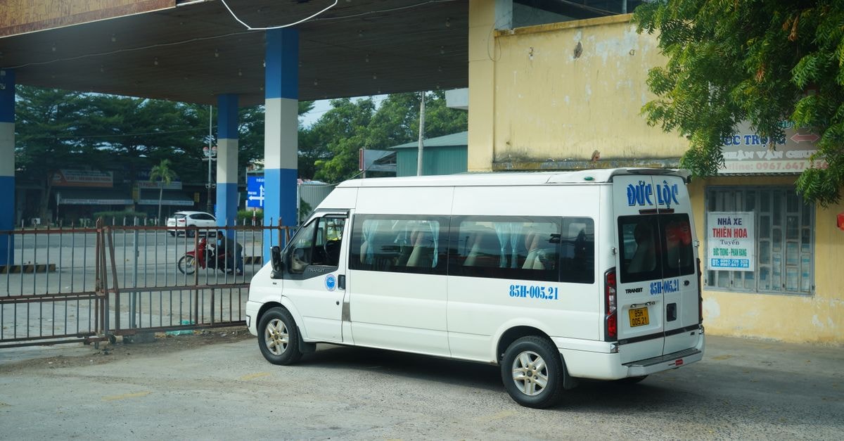 A white minibus is parked at a bus station under a rustic metal canopy.