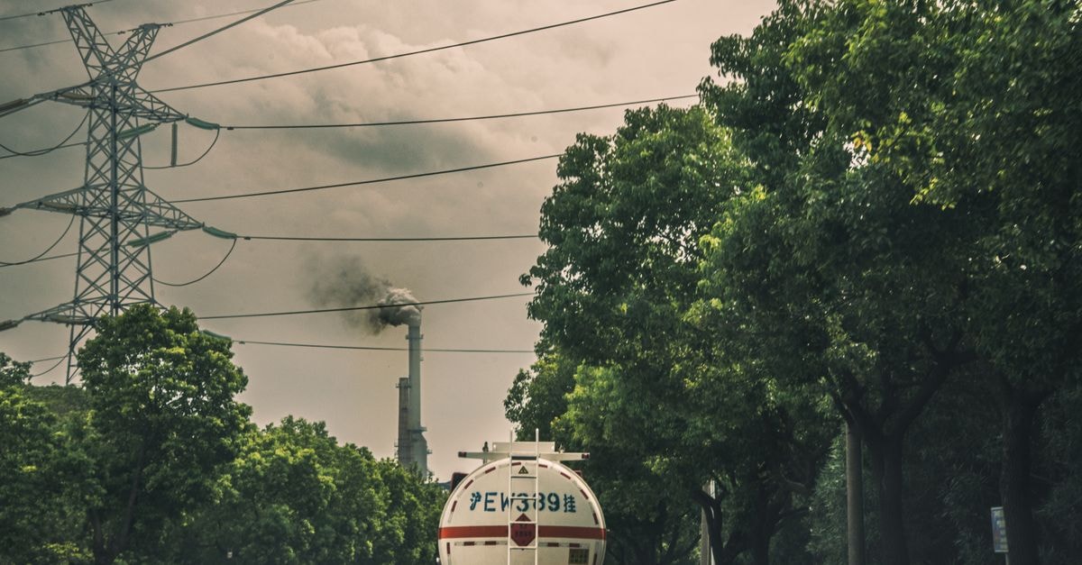 IEA speeds up oil stock release, prioritizes diesel Tanker truck driving on a road by a smokestack emitting smoke under power lines and trees.