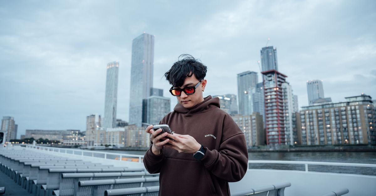 Apple pushes back smart glasses debut as rivals expand wearable bets Casual young man in sunglasses using smartphone on a riverboat in London with city skyline background.