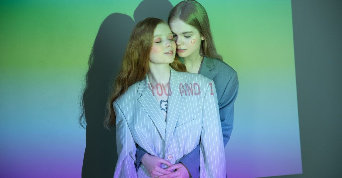 Two women embracing in a studio with colorful light projections and text.