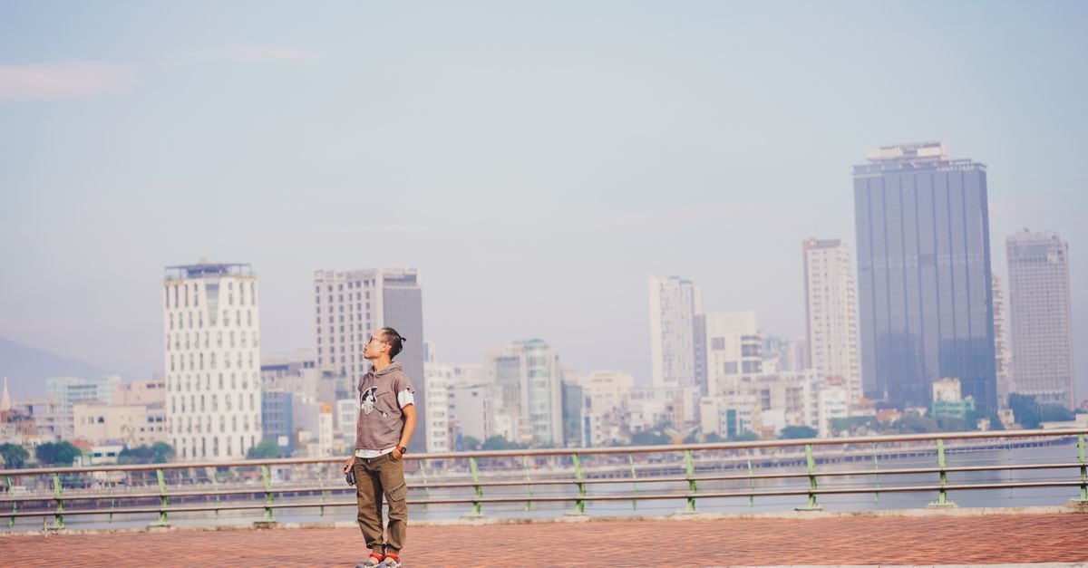 A man stands by a waterfront railing, gazing at a vibrant city skyline under a blue sky.
