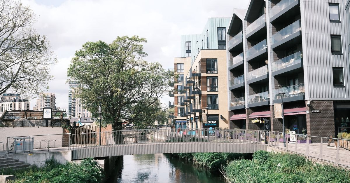 Contemporary apartment buildings line a calm river with a pedestrian bridge in the foreground.