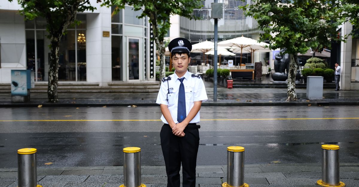 A security officer stands on a wet street in Shanghai after rain, with urban greenery and buildings.