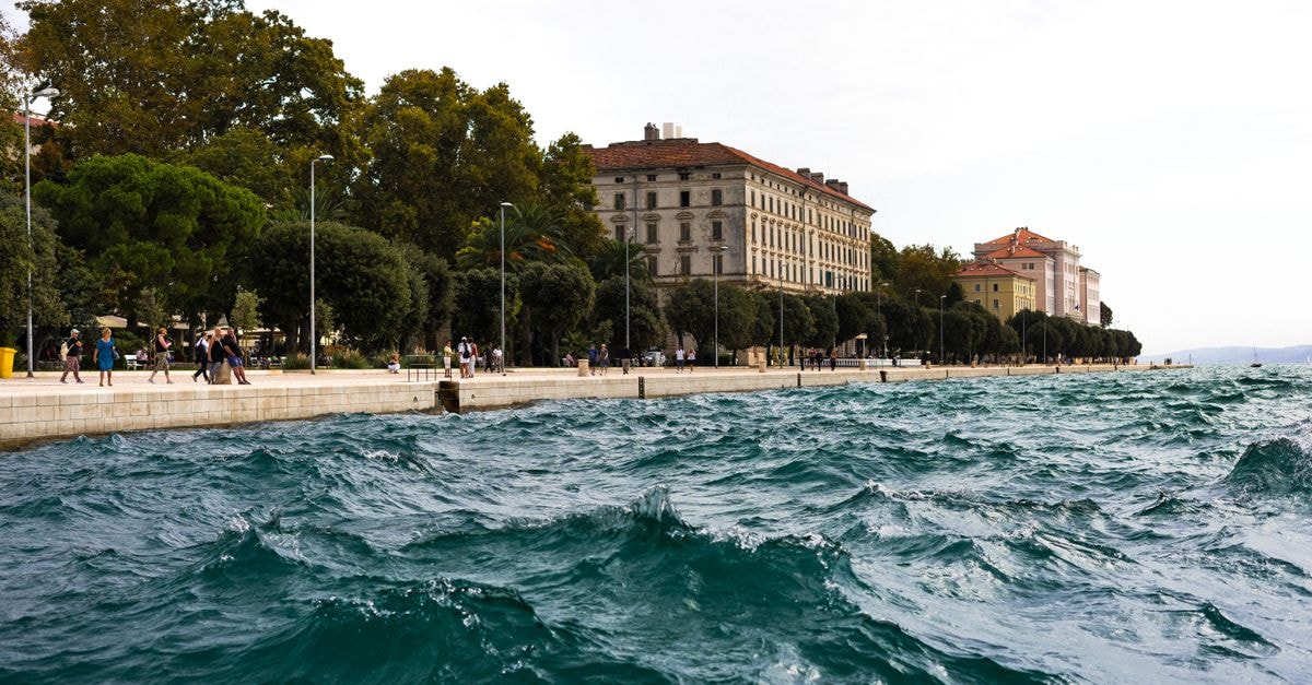 Waves crashing against a scenic coastal promenade lined with trees and historic buildings.