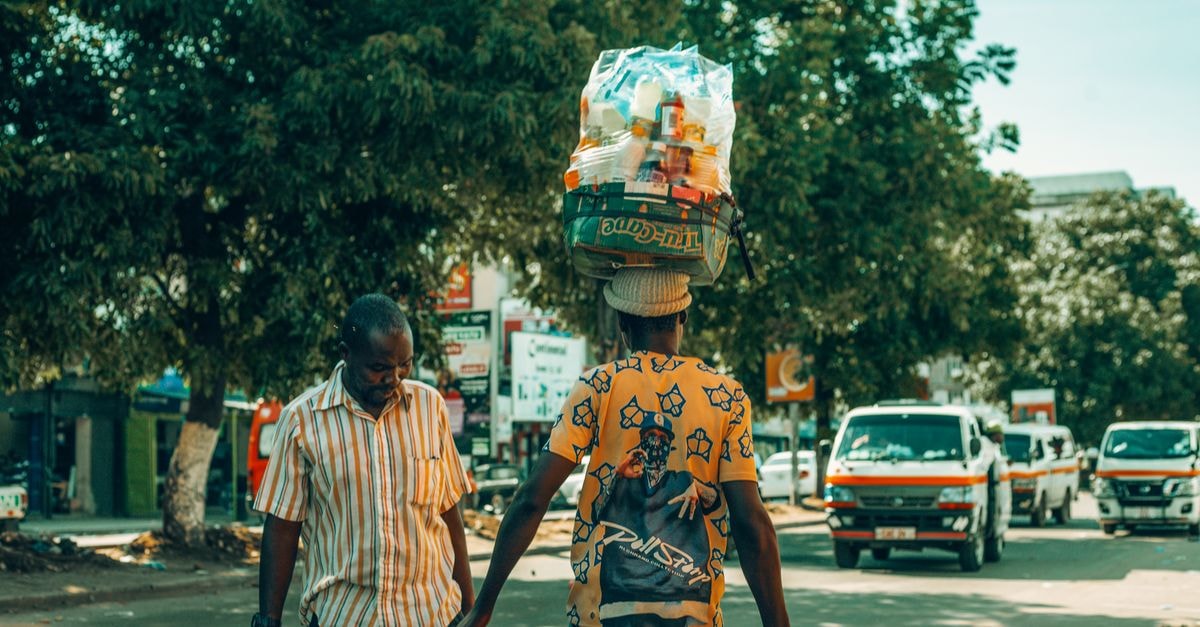 Two men carrying goods through a lively city street on a sunny day.