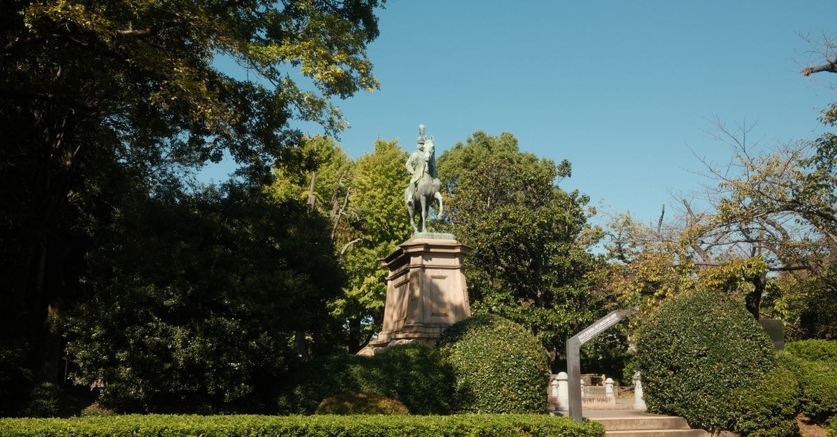 Bronze equestrian statue surrounded by lush greenery under a bright blue sky in Taito City, Tokyo.