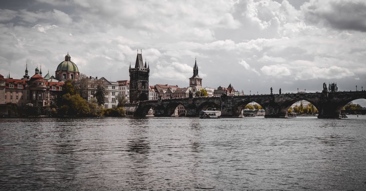 Prague in July 2026: A Complete Summer Travel Guide Iconic view of Charles Bridge spanning Vltava River with Prague's skyline, cloudy skies.
