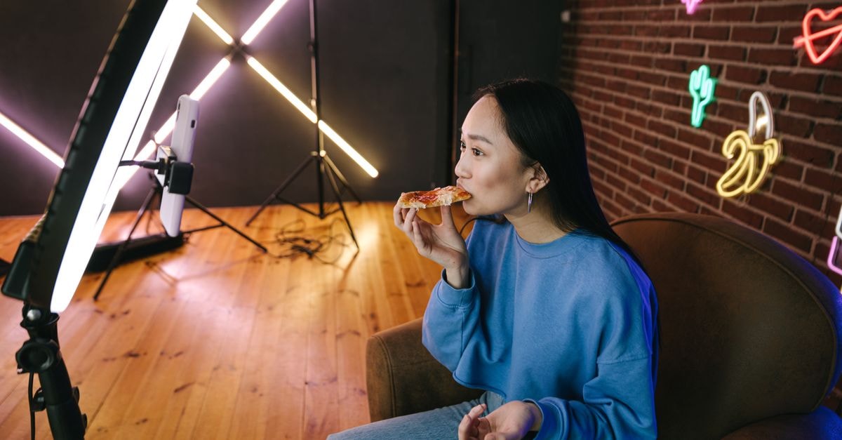 Competitive Eater and 'Amazing Race' Alum Tim Janus Dies at 49 Asian woman eating pizza while streaming indoors with neon lights and ring light setup.