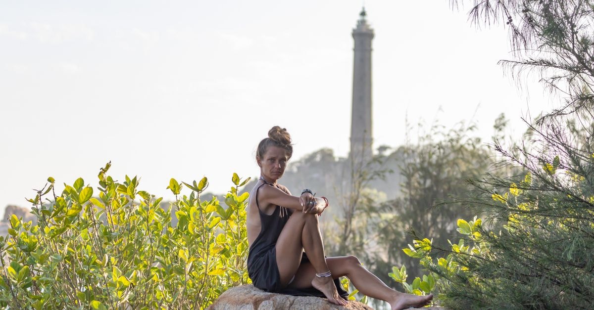 Mahajanga Travel Guide: Harbor Light, Baobab Skies, and Slow Coast Living A woman in casual attire sits on rocks near a lighthouse surrounded by greenery.